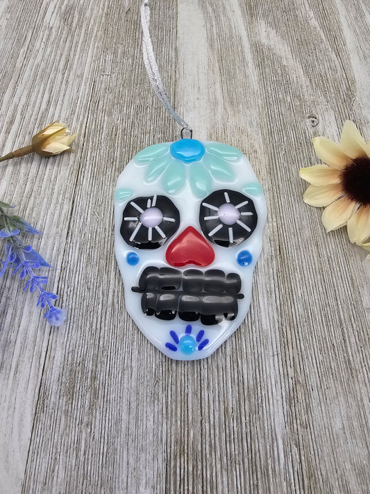 Sugar skull ornament, large black eyes and teeth, heart shape nose.  Blue flower crown and blue dot cheeks accent.