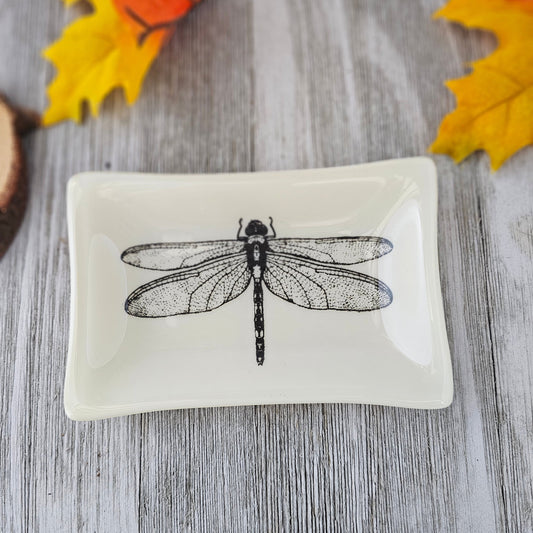 A 4x6 dish, cream colored with a black outline of a dragonfly fused into the glass to be one solid piece.