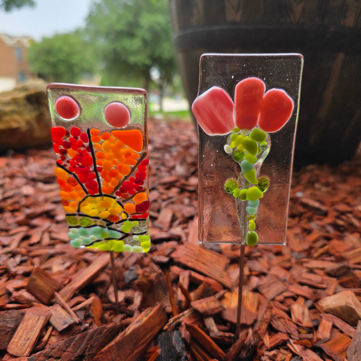 Two garden stakes in a cedar mulched flower bed. One garden stake is a sunset and the other a flower.