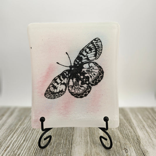 White fused glass dish with a touch of pink and a butterfly motif.  Dish is on a display holder sitting on a gray surface.