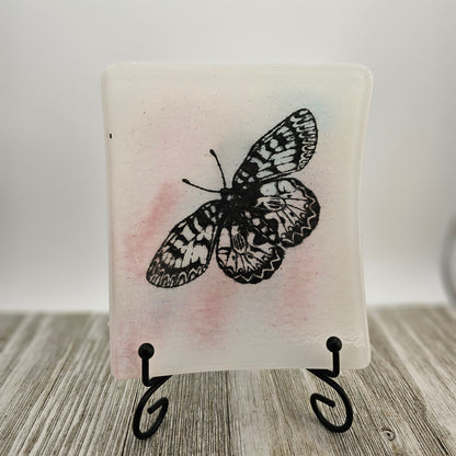 White fused glass dish with a touch of pink and a butterfly motif.  Dish is on a display holder sitting on a gray surface.