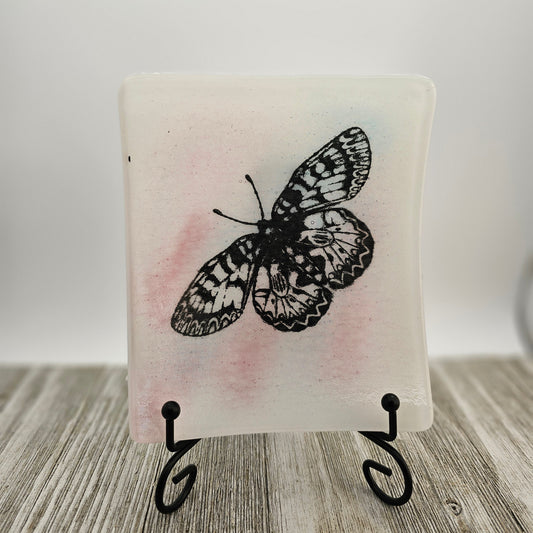 White fused glass dish with a touch of pink and a butterfly motif.  Dish is on a display holder sitting on a gray surface.