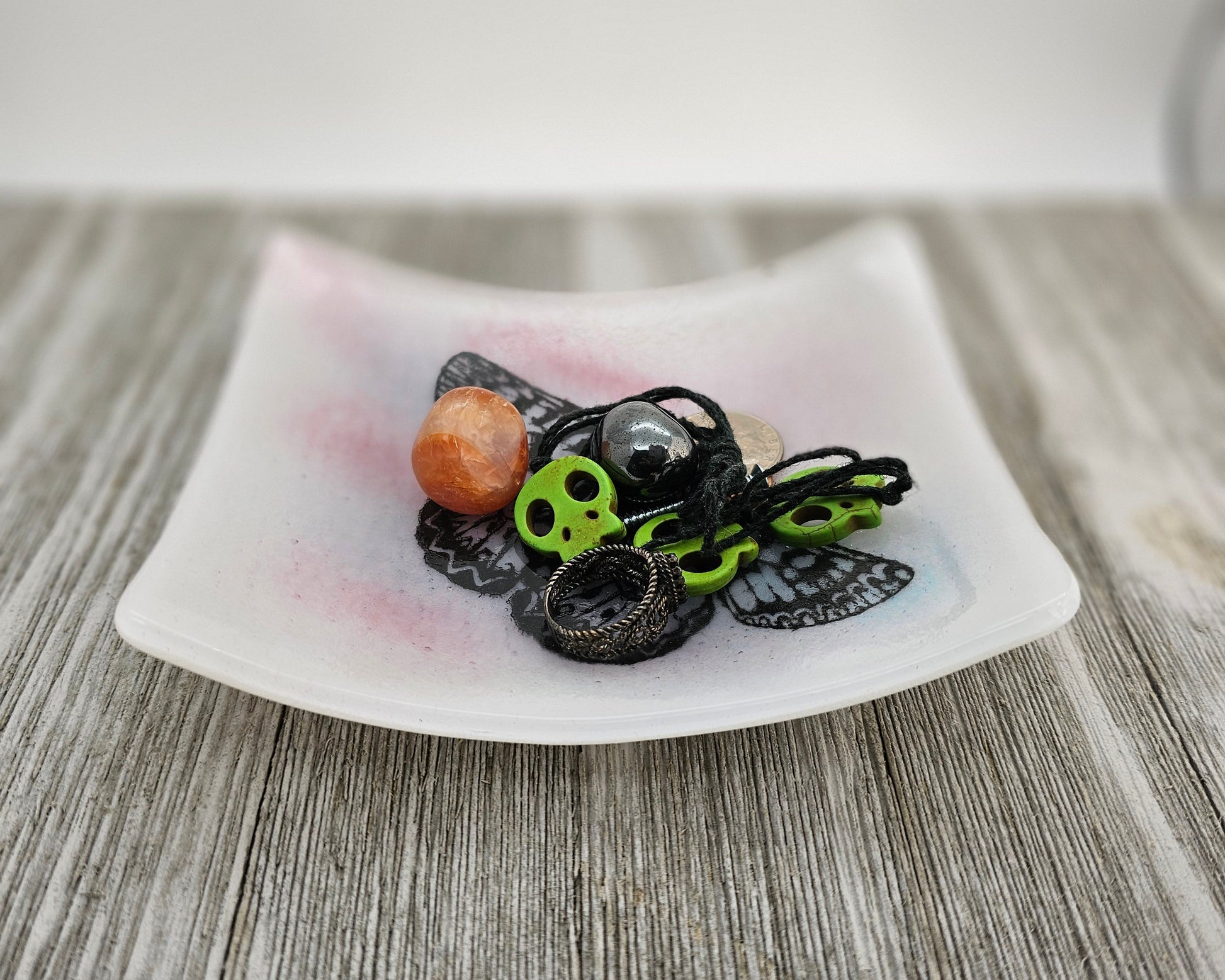 White fused glass dish with a touch of pink and blue and a butterfly motif.  Dish is sitting on a gray surface and is holding trinkets, suggesting use as a trinket holder.