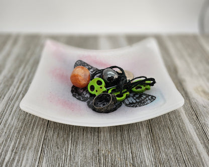 White fused glass dish with a touch of pink and blue and a butterfly motif.  Dish is sitting on a gray surface and is holding trinkets, suggesting use as a trinket holder.