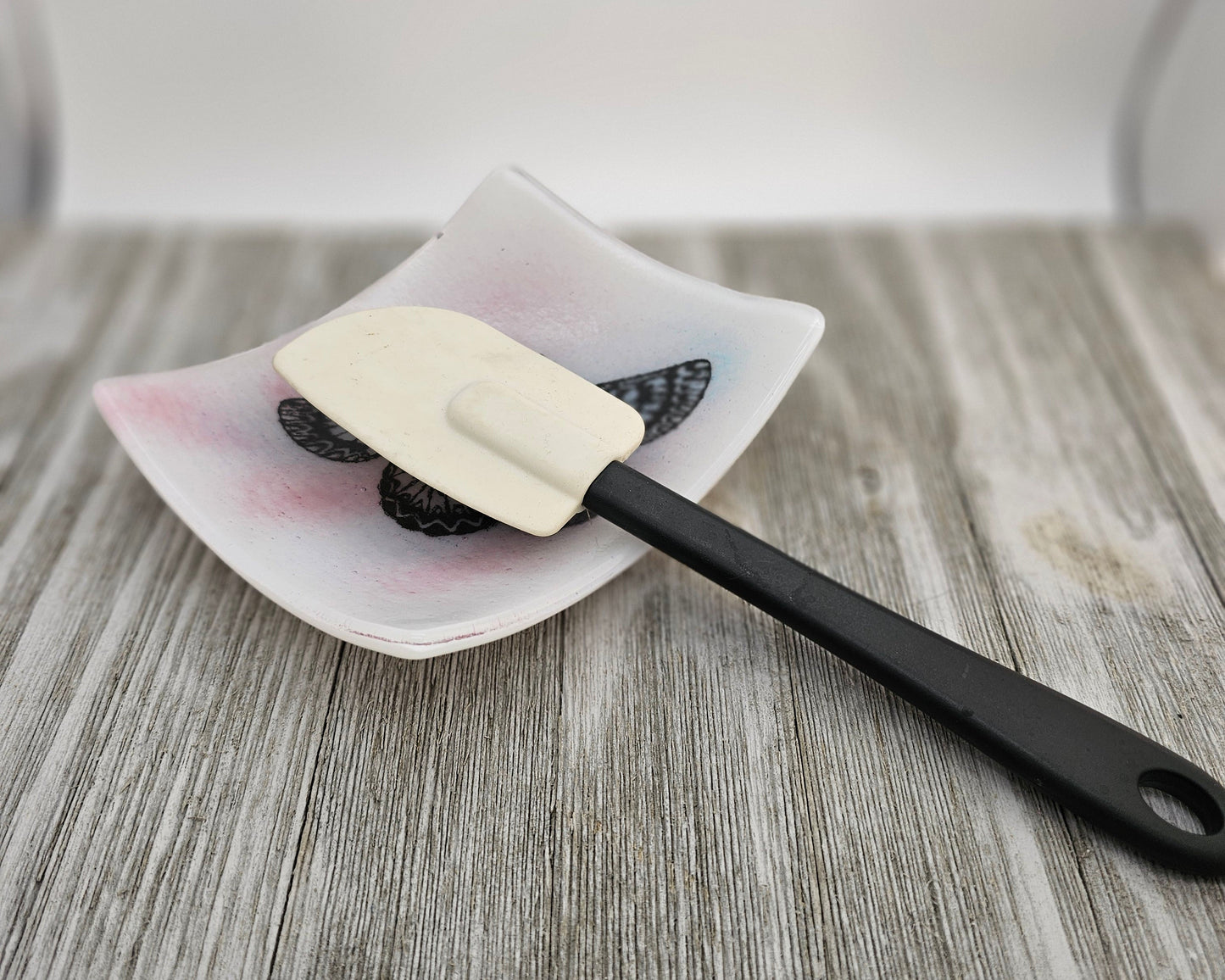 White fused glass dish with a touch of pink and blue and a butterfly motif.  Dish is sitting on a gray surface and is holding a spoon, suggesting use as a spoon holder.