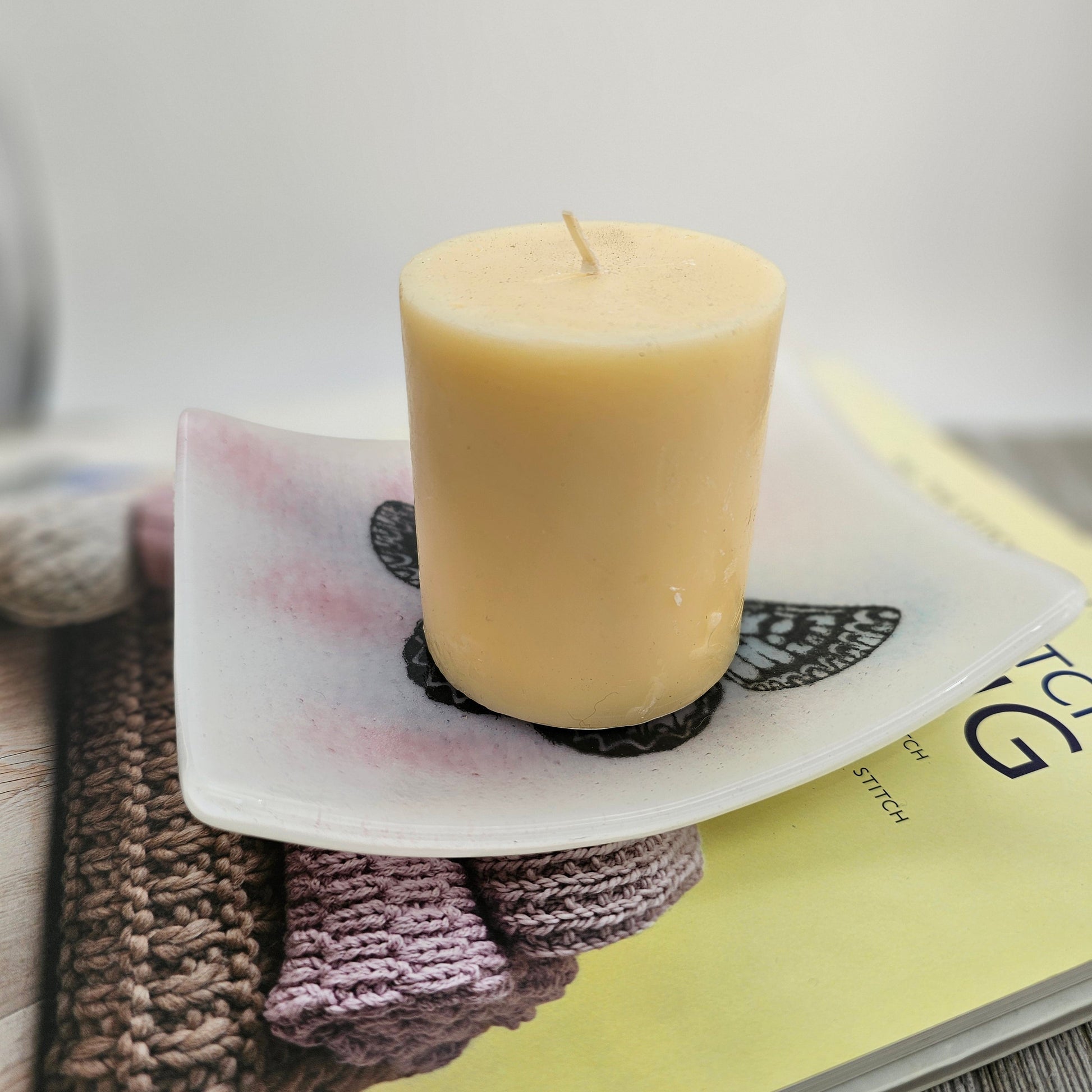 White fused glass dish with a touch of pink and blue and a butterfly motif.  Dish is sitting on a book on a gray surface and is holding a large candle, suggesting use as a candle holder.