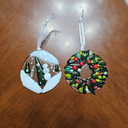 pair of fused glass ornaments. One depicts a snowing scene in the woods with a snowman. The other a wreath with berries.