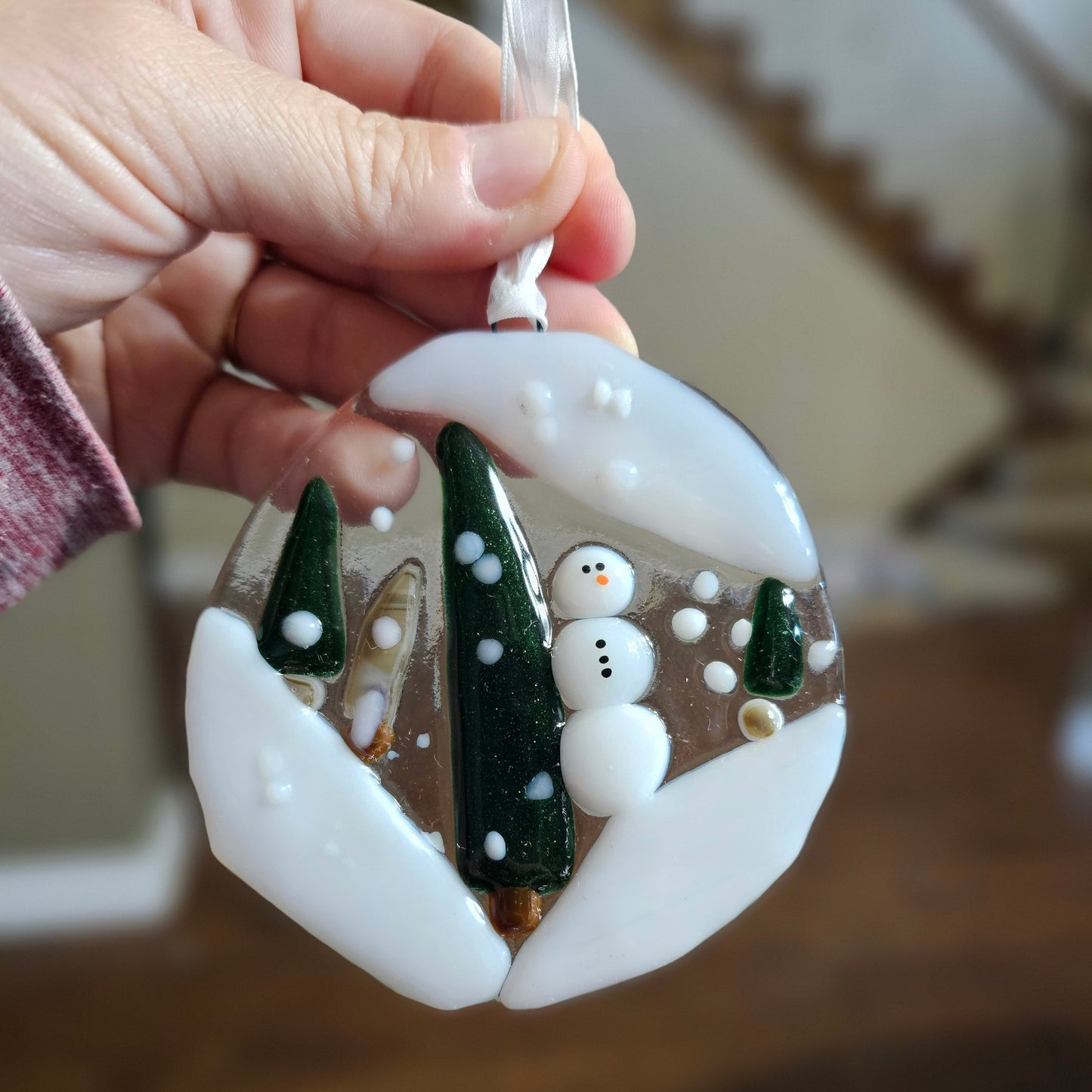 A hand holding an ornament depicting a snowy woods with a snowman.  The ornament is glass