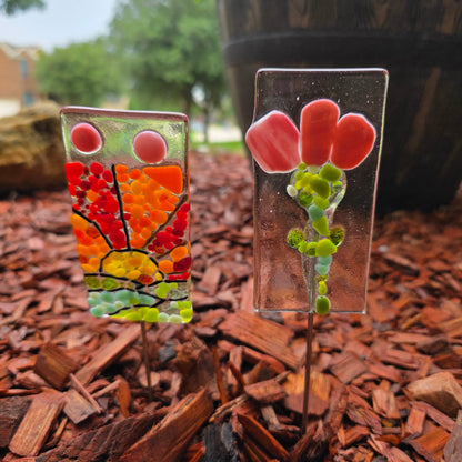 Two garden stakes in a cedar mulched flower bed.  One garden stake is a sunset and the other a flower.