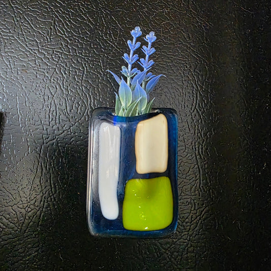 Small magnet vase with silk flowers on the side of a black fridge.