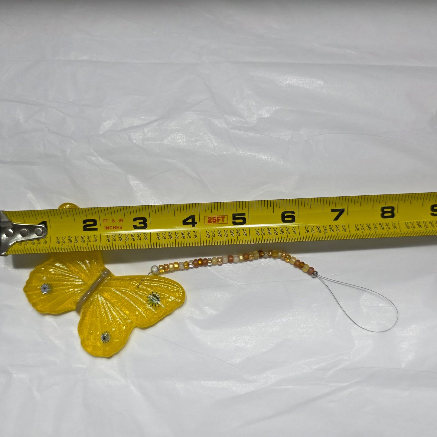 Yellow butterfly hanging on a beaded wire with a white background against a measure tape to show approximate hang length. About 8.5 inches.