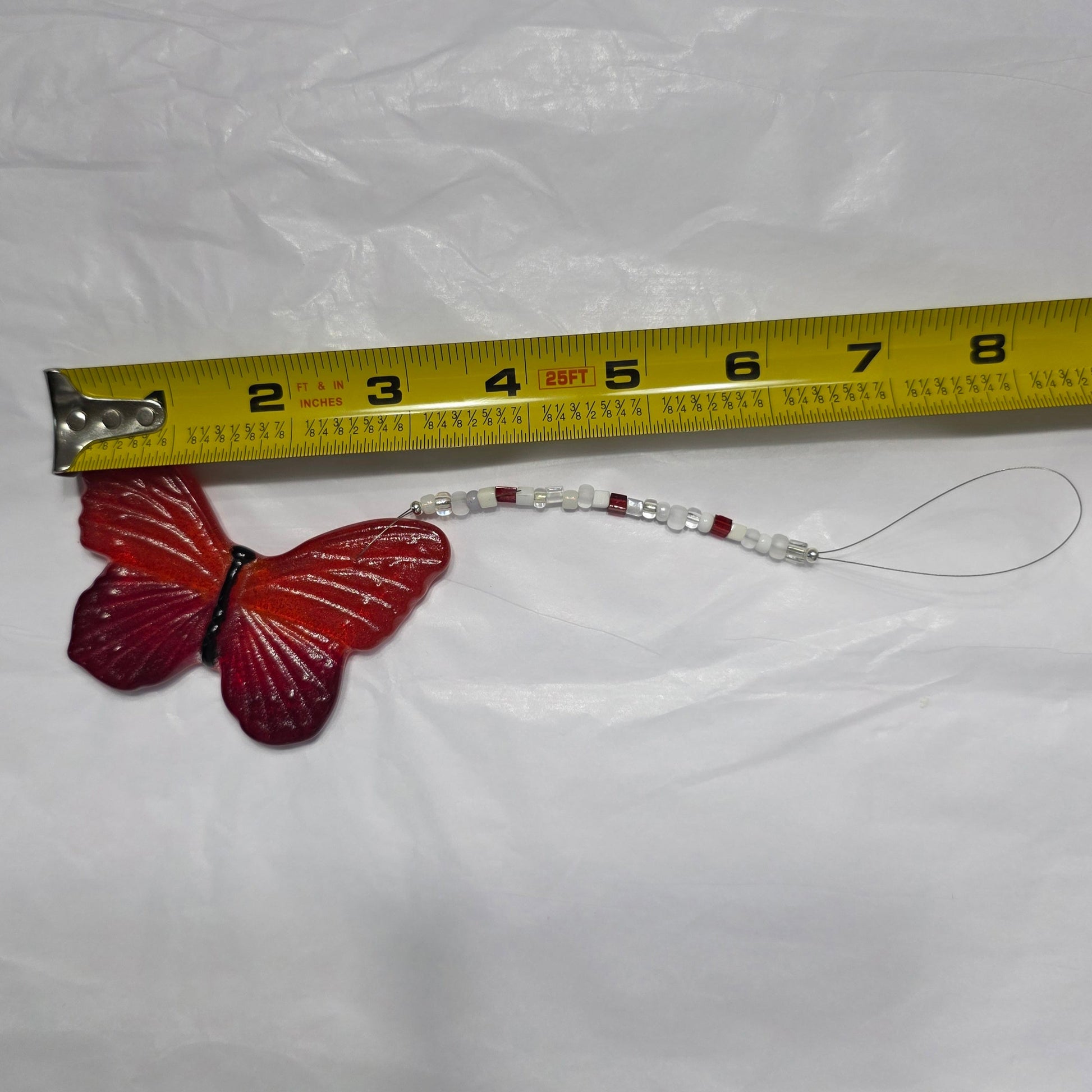 Hanging by  a beaded wire this red and orange butterfly is being measured for its approximate hang length; 8.5 inches