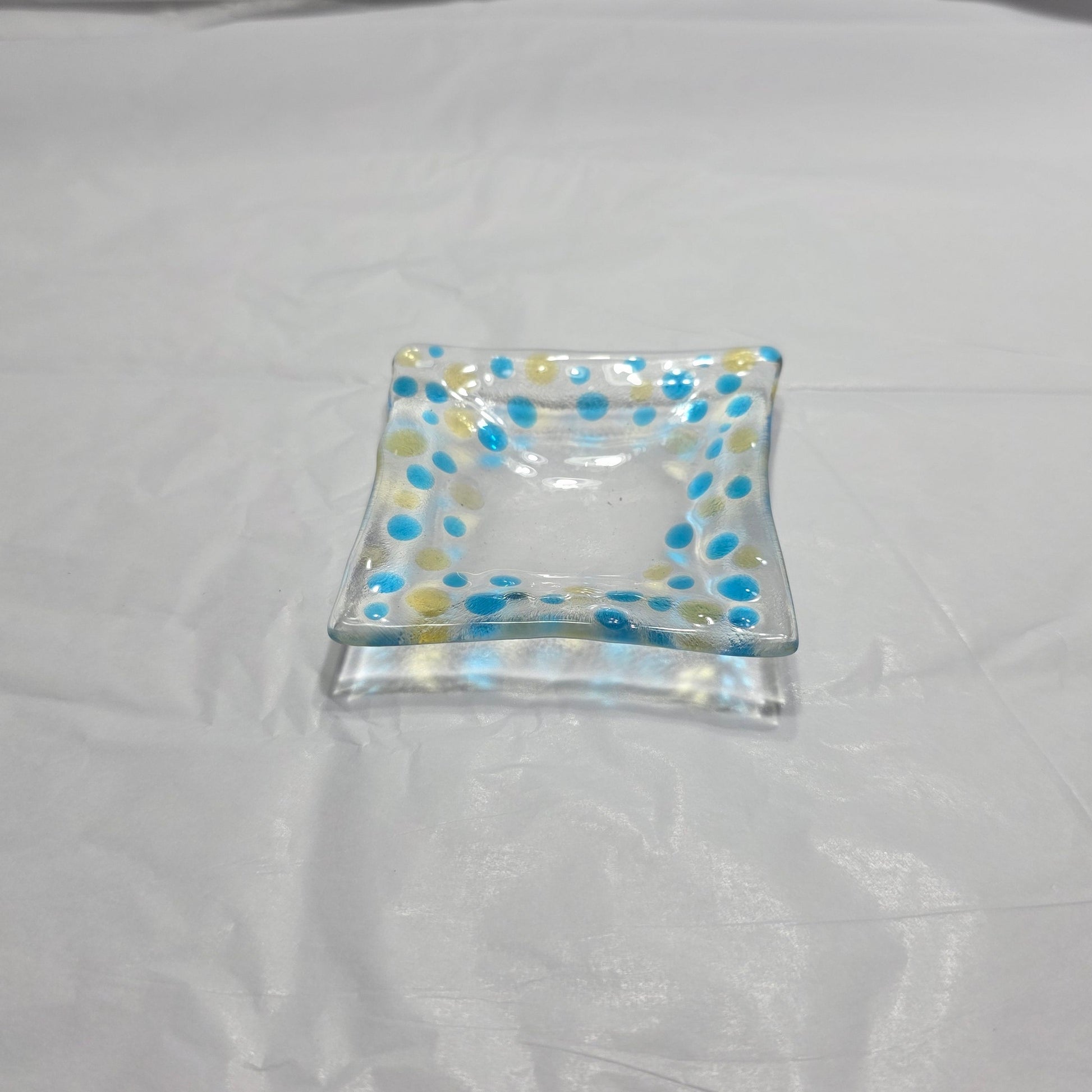 A clear dish with blues and yellow polka dots, sitting on a white surface. The colored dots reflect on the white surface from the rooms light source.