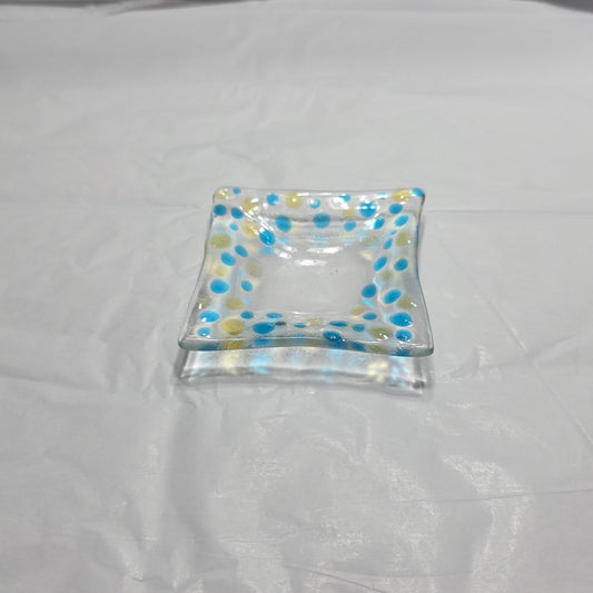 A clear dish with blues and yellow polka dots, sitting on a white surface. The colored dots reflect on the white surface from the rooms light source.