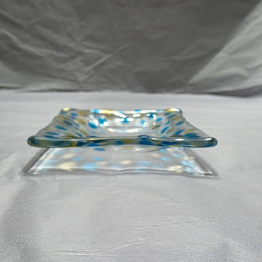 A clear dish with blues and yellow polka dots, sitting on a white surface. The colored dots reflect on the white surface from the rooms light source.