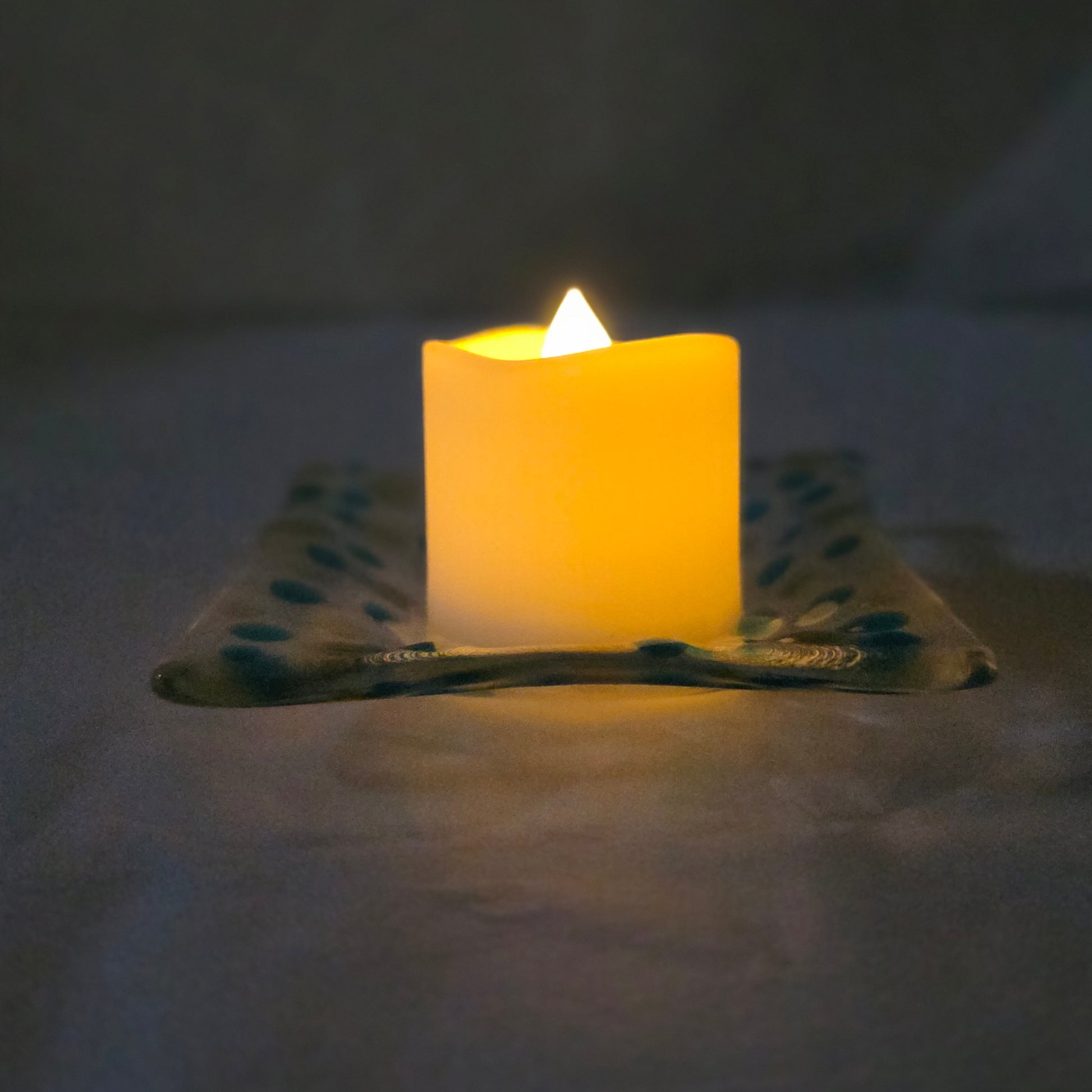Side view of a clear dish with blues and yellow polka dots, sitting on a white surface.  The dish has a battery-operated candle displayed on it, suggesting a use as a candle holder.
