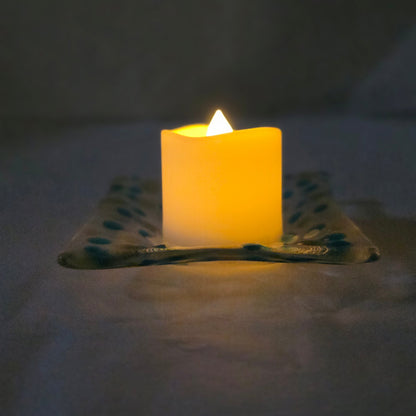 Side view of a clear dish with blues and yellow polka dots, sitting on a white surface.  The dish has a battery-operated candle displayed on it, suggesting a use as a candle holder.
