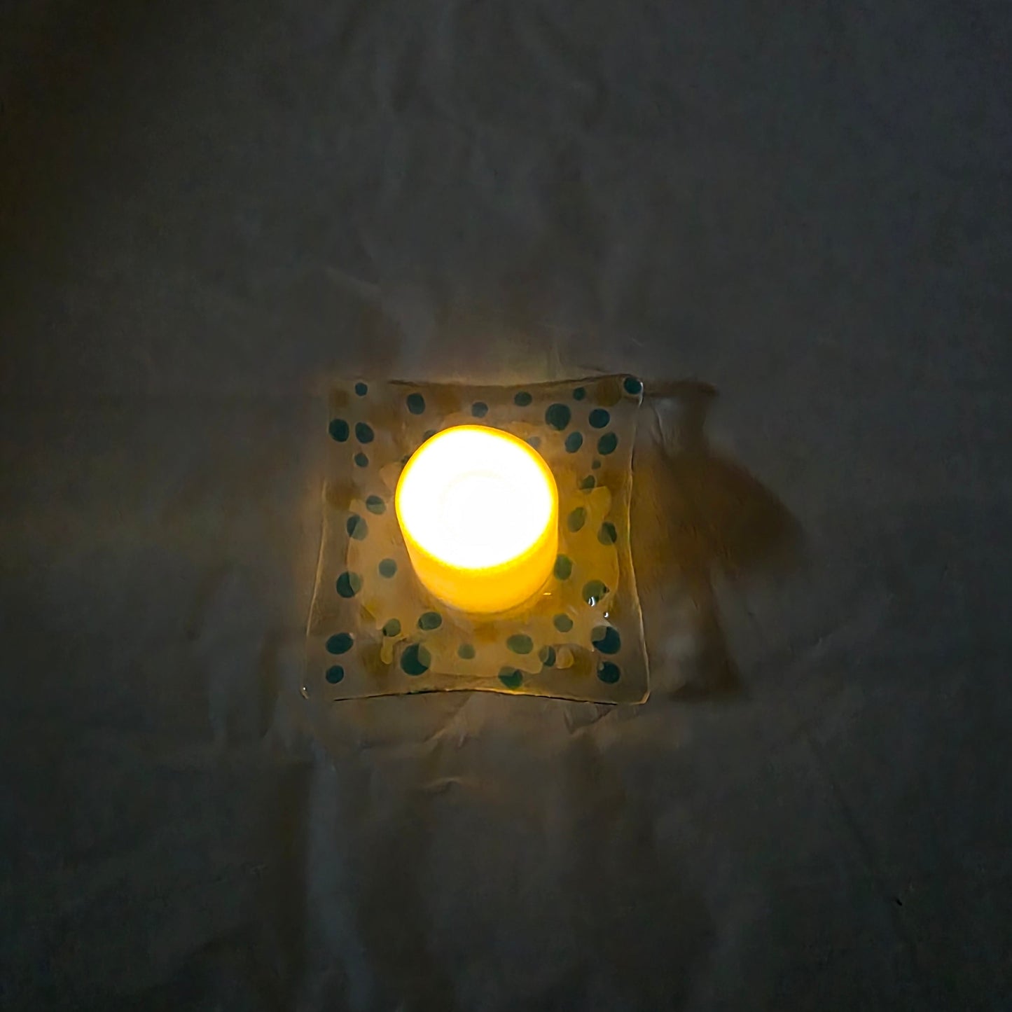 A top-down view of a clear dish with blues and yellow polka dots, sitting on a white surface.  The dish has a battery-operated candle displayed on it, suggesting a use as a candle holder.