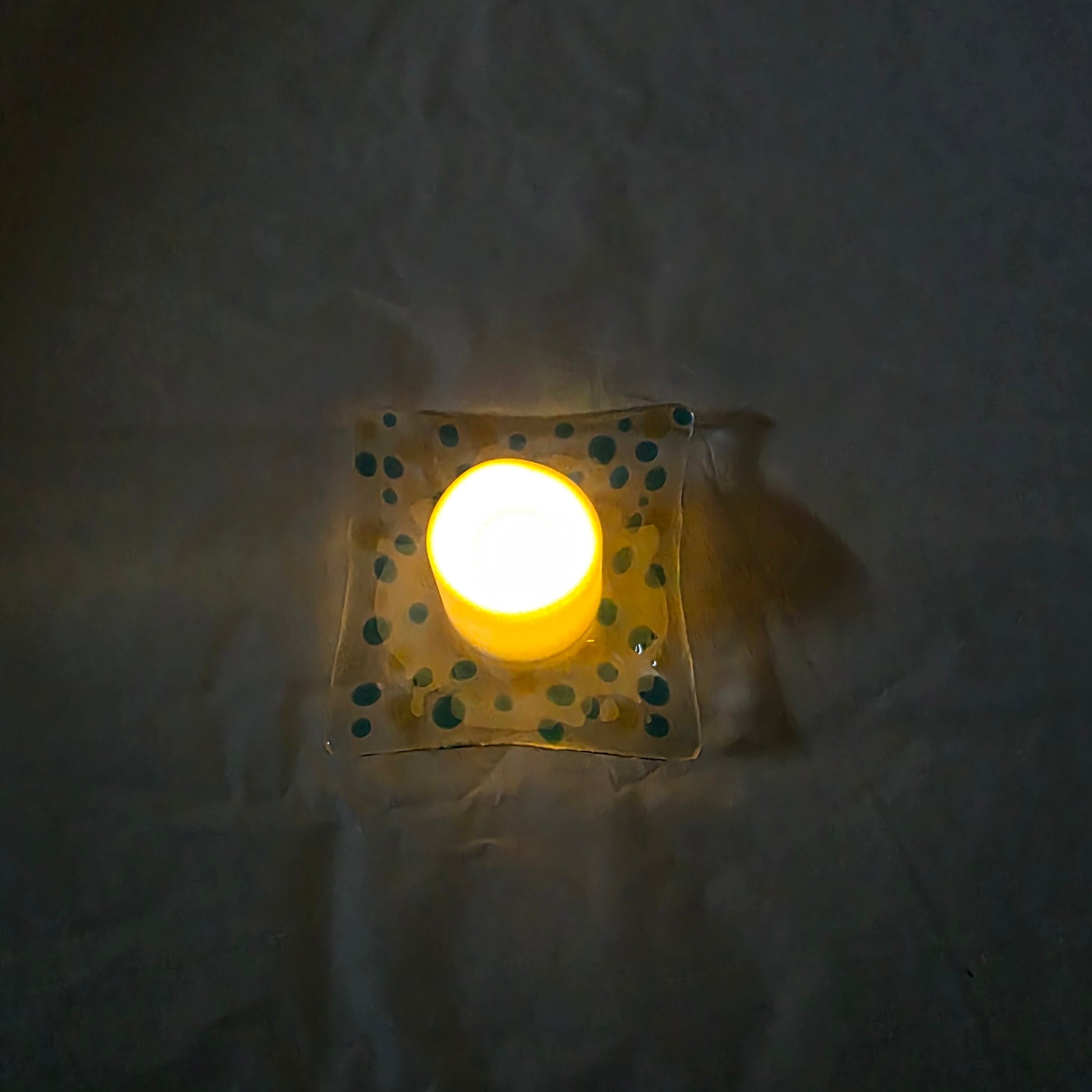 A top-down view of a clear dish with blues and yellow polka dots, sitting on a white surface.  The dish has a battery-operated candle displayed on it, suggesting a use as a candle holder.