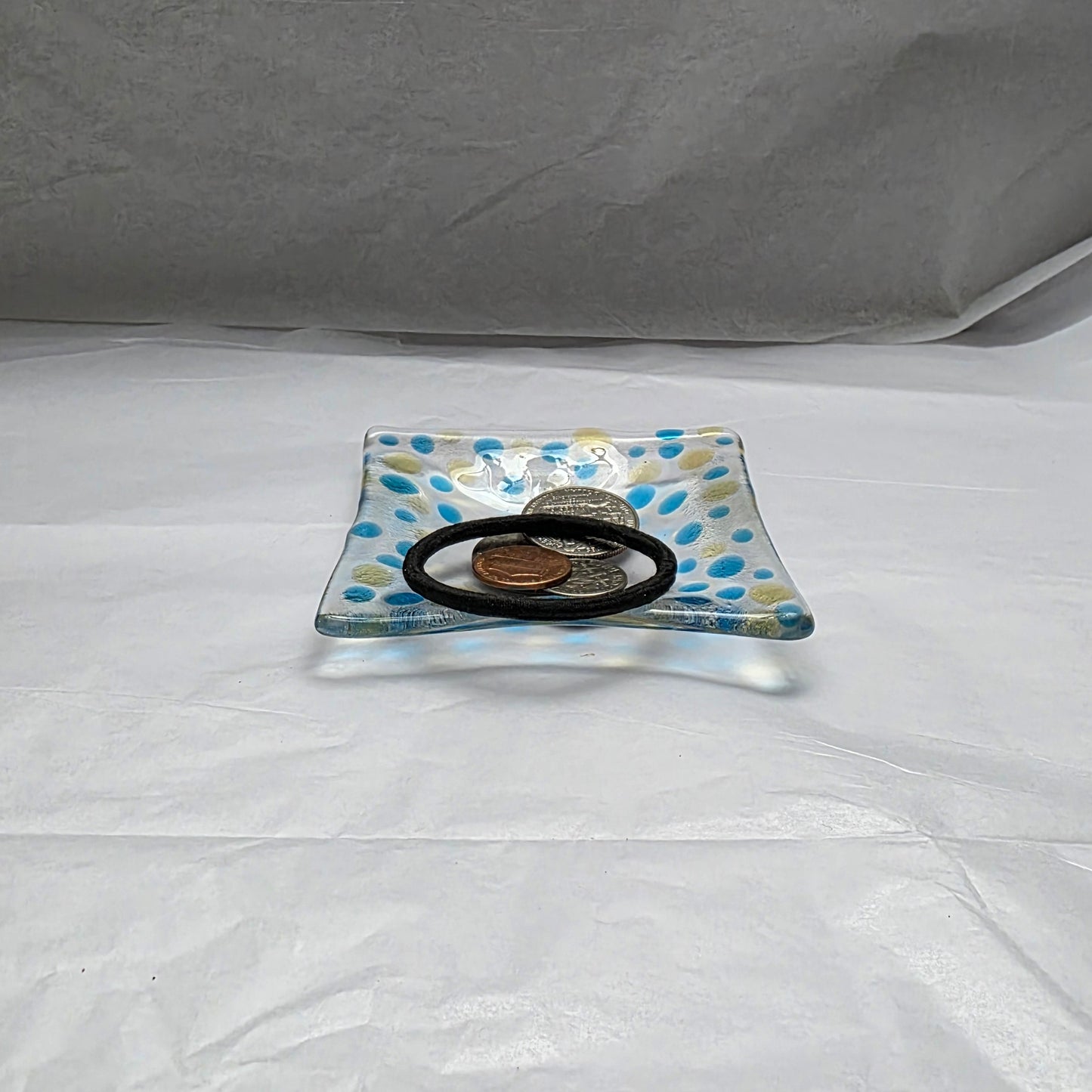 A clear dish with blues and yellow polka dots, sitting on a white surface.  The dish has coins and a hair tie displayed on it, suggesting a use as a catch-all or trinket dish.