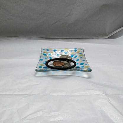 A clear dish with blues and yellow polka dots, sitting on a white surface.  The dish has coins and a hair tie displayed on it, suggesting a use as a catch-all or trinket dish.