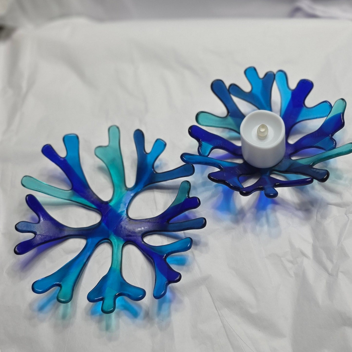 Two bowls of various shade of blue glass candle holder in a coral reef design. The candle holder has a white LED candle in the center. The candle holder is on a wooden surface with seashells.