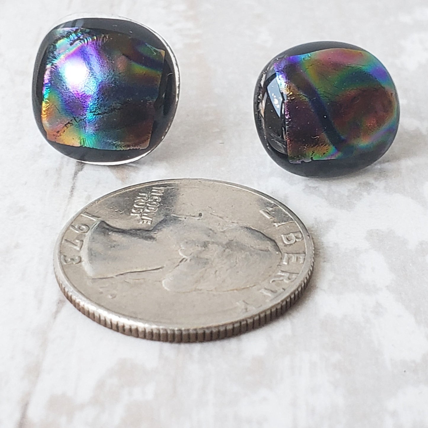 Black stud earrings with a rainbow iridescent top and silver-plated posts.  The earrings are being compared to the size of a quarter.